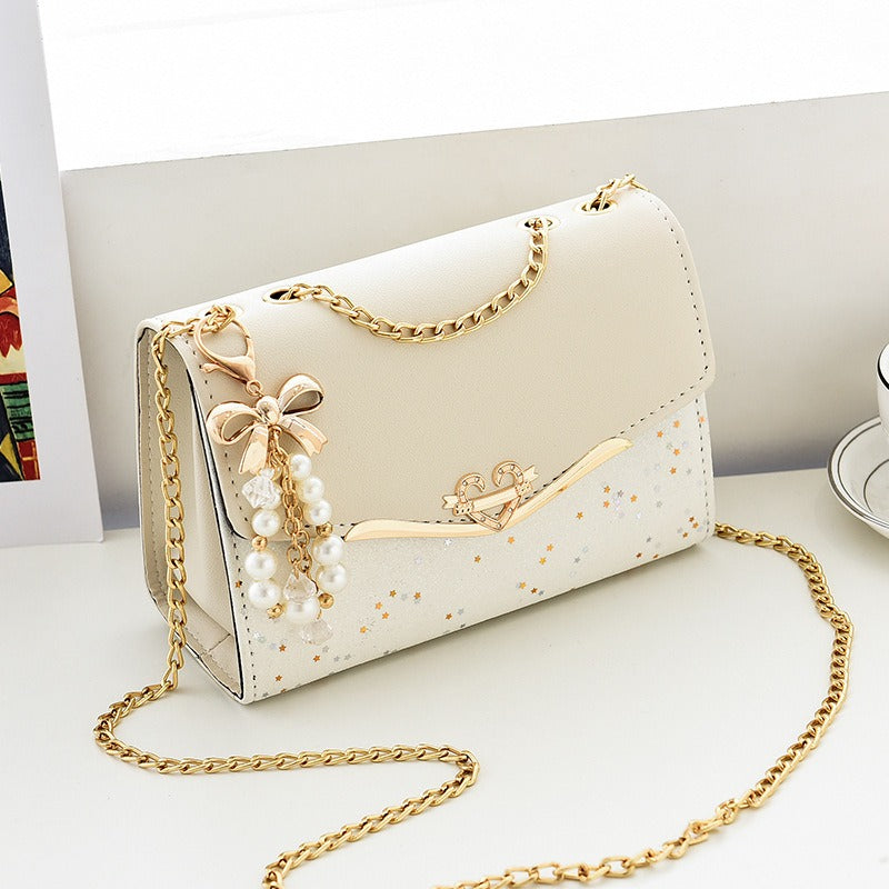 Sac cérémonie blanc à chaîne dorée, charm perlé et finition pailletée, format 21x14x8 cm, posé sur fond blanc

